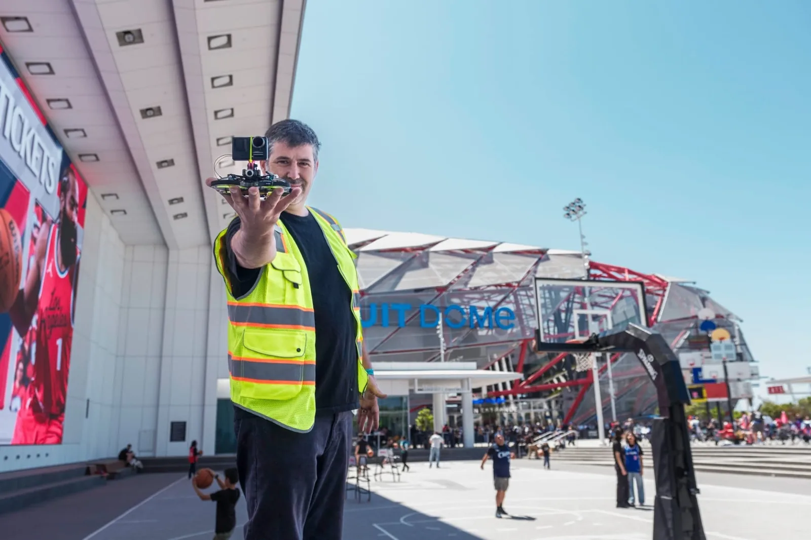 Jason El Moussa in a hi-vis safety vest holding a ducted FPV cinewhoop with an action camera mounted on top, on an outdoor court in front of an arena with a crowd behind him